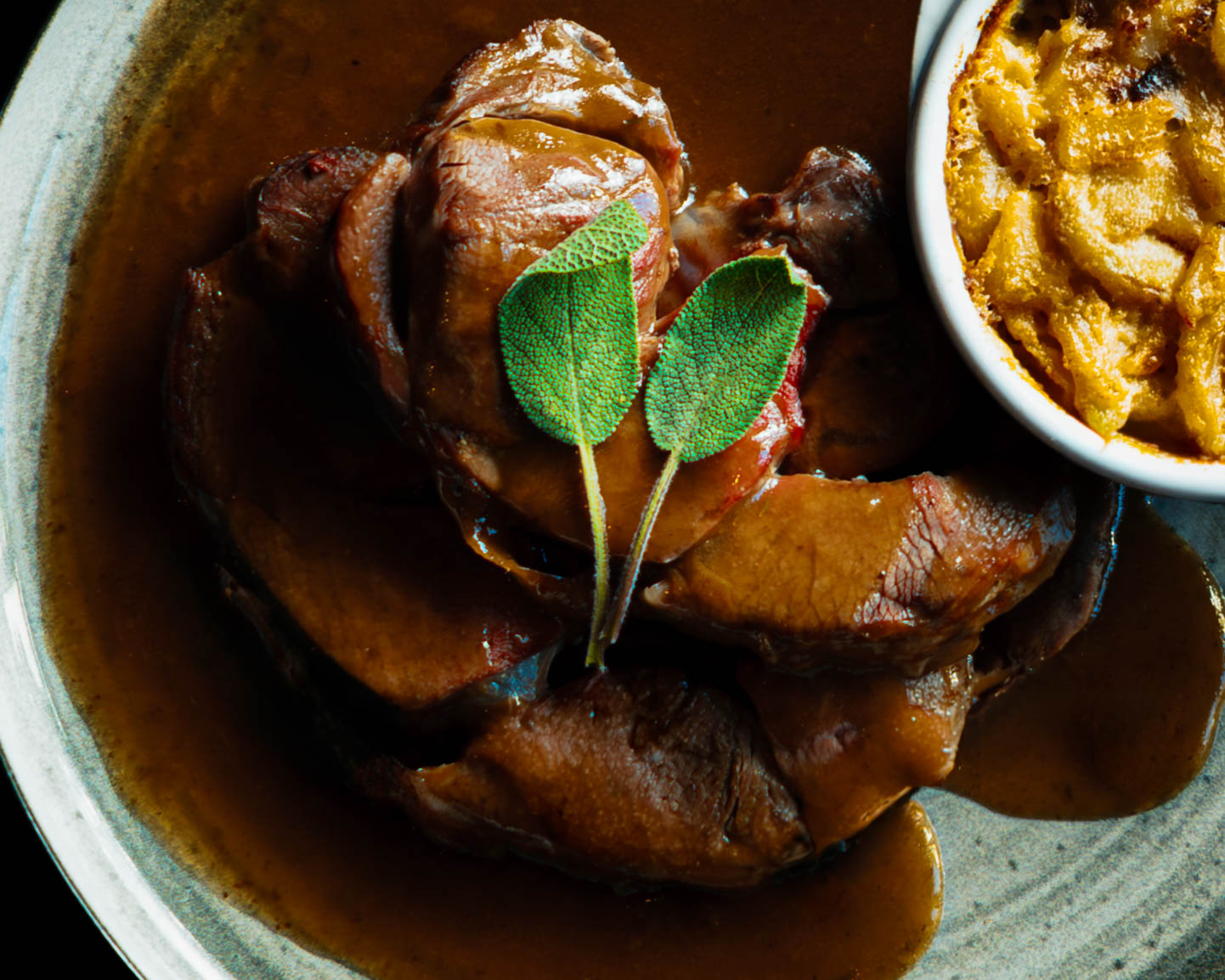 Gros plan d’un plat de viande nappée de sauce, feuilles de sauge et accompagnement gratiné